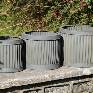 May include: Three grey, ribbed, barrel-shaped planters of different sizes. The planters have a circular rim and base. They are arranged on a stone surface with greenery in the background. Suitable for outdoor use, these planters are ideal for gardens.