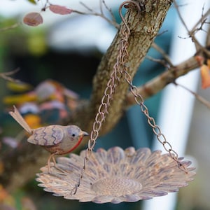 May include: A rustic metal bird feeder with a sunflower design, suspended by a chain from a tree branch. A small, detailed metal bird with red accents perches nearby. The feeder has a weathered, antique finish.