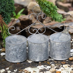 Op de afbeelding: Een gegalvaniseerde metalen plantenbak met drie cilindrische potten. De potten zijn verbonden door een decoratief frame in smeedijzeren stijl met krullen. De plantenbak is ontworpen voor buitengebruik en is geschikt voor kleine planten of kruiden.