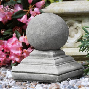 May include: A grey concrete finial featuring a smooth spherical top resting on a tiered square base. The object has a weathered, textured surface and is positioned in an outdoor setting with pink flowers and stone elements visible in the background.