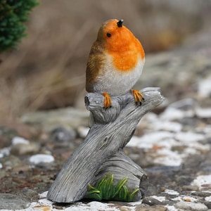 Op de afbeelding: Gedetailleerd beeldje van een roodborstje dat op een verweerde, grijze tak zit. De vogel heeft een feloranje borst, bruine vleugels en een witte buik. De tak heeft groen gras aan de basis, waardoor een naturalistisch tuinornament ontstaat.