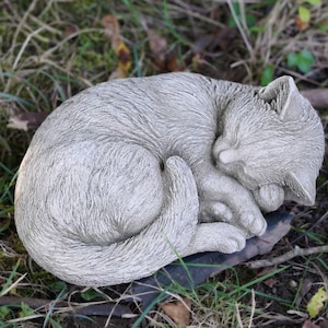 Könnte beinhalten: Graue, schlafende Katzenstatue, zusammengerollt. Das Fell der Katze ist strukturiert, die Pfoten sind unter dem Körper versteckt. Die Statue besteht aus einem steinartigen Material und liegt auf einer dunklen Oberfläche, umgeben von Gras und Blättern.