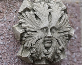 May include: A stone sculpture of a Green Man's face with a leafy beard and hair. The sculpture is carved in a starburst shape and is mounted on a stone wall.