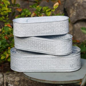 Peut inclure: Un ensemble de trois jardinières ovales en métal avec une finition argentée vieillie. Chaque jardinière présente un motif de volutes en relief répété sur les côtés extérieurs, créant un aspect texturé sur les contenants empilés.
