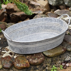 May include: A galvanized metal oval planter with two curved handles. The planter has a ribbed design and is sitting on a bed of rocks. The overall color is a muted gray, with variations in tone due to the metal finish.