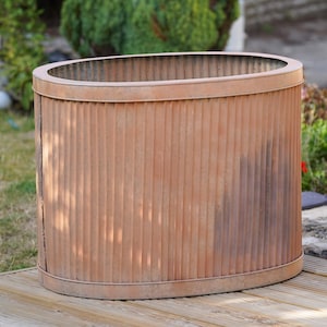 Peut inclure: Une jardinière de forme ovale avec un extérieur en métal ondulé présentant une finition rouillée orange-brun patinée. Le récipient possède un bord plat et lisse en haut et en bas, avec des nervures verticales sur toute la longueur du corps.