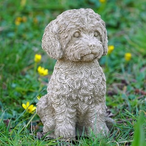 Puede incluir: Una estatua de perro de color piedra, sentada en un entorno de hierba con flores amarillas. La escultura presenta un pelaje rizado y texturizado. Los rasgos del perro están bien definidos, lo que le da un aspecto realista.