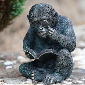 Könnte beinhalten: Bronze-farbene Schimpansen-Statue, sitzend und lesend in einem Buch mit Monokel. Die Skulptur hat eine verwitterte Oberfläche, mit Details im Fell und Buch. Ein dekorativer Artikel für den Innen- oder Außenbereich.