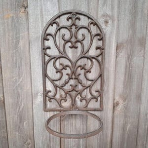 Może przedstawiać: Brązowy, metalowy uchwyt na donicę montowany na ścianie. Uchwyt ma łukowaty wierzch z ozdobnym, symetrycznym wzorem z zawijasów. Poniżej umieszczono okrągły pierścień do trzymania donicy. Zamontowany na drewnianym tle.