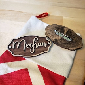 May include: A wooden stocking hanger with the name "Meghan" engraved on it. The hanger is brown and has a decorative design. It comes with a safety pin for attaching it to a stocking.