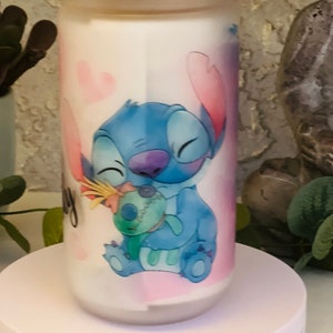 May include: A white ceramic jar with a pink lid and a blue and pink cartoon character design. The character is Stitch from the Disney movie Lilo & Stitch. Stitch is holding a small blue and green character. The jar has a pink and white background with hearts.
