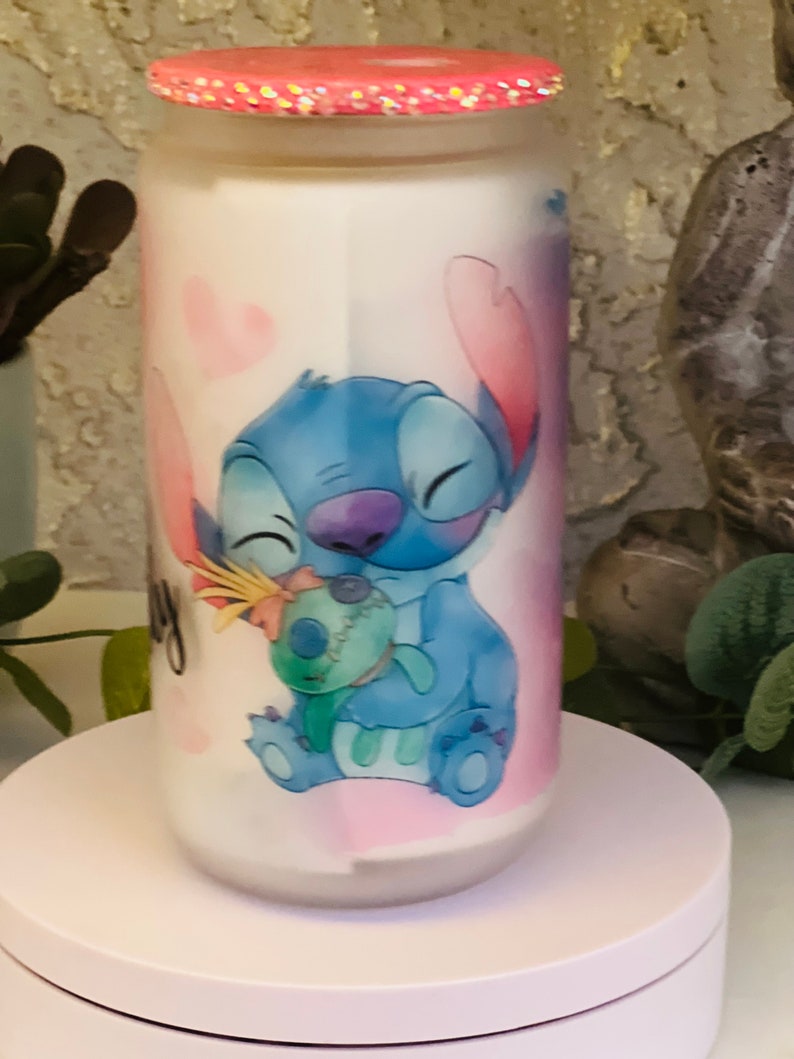 May include: A white ceramic jar with a pink lid and a blue and pink cartoon character design. The character is Stitch from the Disney movie Lilo & Stitch. Stitch is holding a small blue and green character. The jar has a pink and white background with hearts.