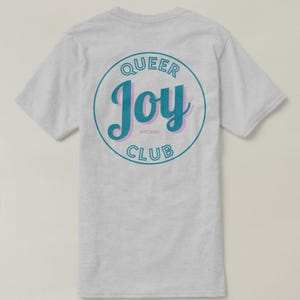 May include: Light gray t-shirt with a circular design on the back. The design features the words "QUEER JOY CLUB" in teal and pink, with "EST. 2023" below the word "Joy". The shirt has short sleeves and a crew neck.