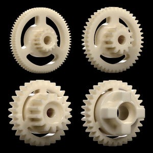 May include: Four off-white plastic gears of varying designs are displayed against a black background. Each gear has a central hub and toothed outer ring. The gears are likely components for machinery or mechanical devices.