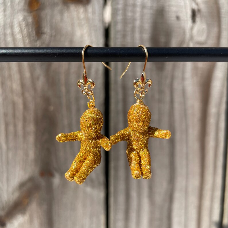 May include: Gold glitter dangle earrings featuring a small human figure with a fleur-de-lis charm at the top of each earring.