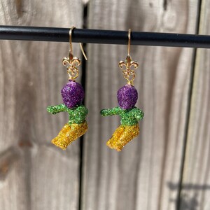 May include: A pair of gold dangle earrings featuring a purple, green, and gold glitter figure with a fleur-de-lis charm.