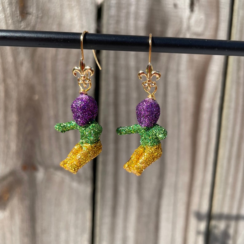 May include: A pair of gold dangle earrings featuring a purple, green, and gold glitter figure with a fleur-de-lis charm.