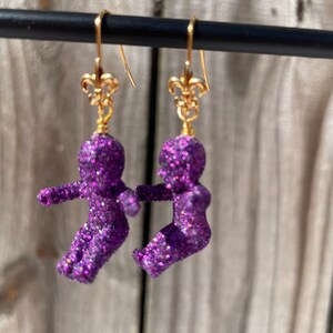 May include: Purple glitter doll earrings with gold fleur-de-lis charms. The dolls are in a standing position with their arms outstretched.