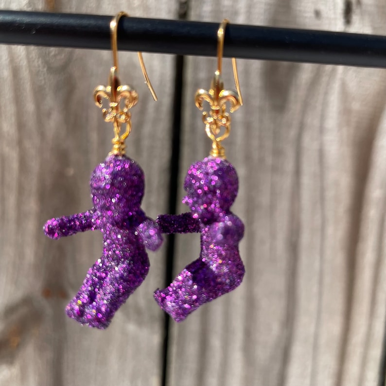 May include: Purple glitter doll earrings with gold fleur-de-lis charms. The dolls are in a standing position with their arms outstretched.