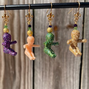 May include: Three pairs of dangle earrings featuring a small glitter baby doll charm. The charms are purple, nude, and green. Each earring has a gold fleur-de-lis charm and a gold hook.