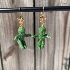 May include: A pair of green glitter earrings with gold fleur-de-lis charms. The earrings are shaped like small people.
