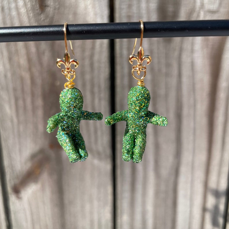 May include: A pair of green glitter earrings with gold fleur-de-lis charms. The earrings are shaped like small people.
