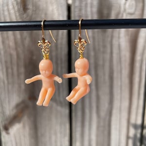 May include: A pair of gold earrings with a small doll figure hanging from each. The dolls are nude and have a light pink color. The dolls are suspended from a gold fleur-de-lis charm.