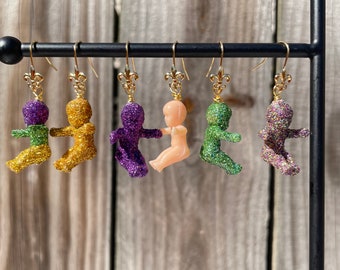 May include: Six pairs of dangle earrings featuring a glittery baby doll charm. The charms are in various colours, including green, gold, purple, and pink.