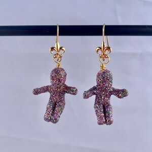 May include: A pair of gold dangle earrings with a purple glitter gingerbread person charm. The charms are detailed with a gold fleur-de-lis at the top.