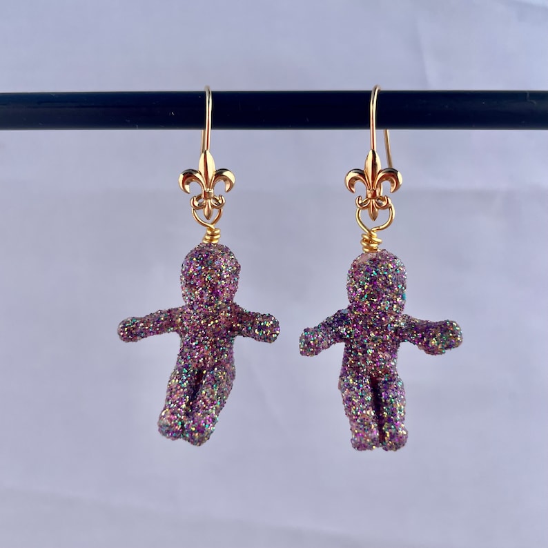 May include: A pair of gold dangle earrings with a purple glitter gingerbread person charm. The charms are detailed with a gold fleur-de-lis at the top.