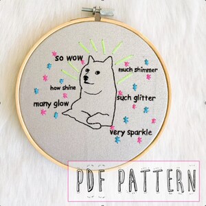 May include: A white dog with a surprised expression is embroidered on a gray fabric in a wooden embroidery hoop. The dog is surrounded by colorful stars and the text "so wow", "much shimmer", "how shine", "many glow", "such glitter", and "very sparkle".