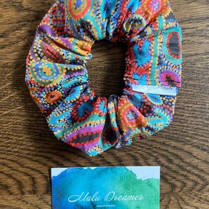 May include: A fabric scrunchie featuring a vibrant dot-art pattern in shades of orange, blue, purple, and black. A small white tag reads Emu Dreaming. Below, a rectangular card displays a watercolor wash and the text Malu Dreamer Handcrafted on Darkinjung Land.