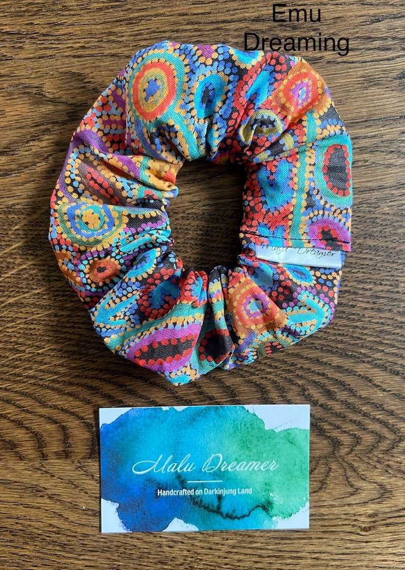 May include: A fabric scrunchie featuring a vibrant dot-art pattern in shades of orange, blue, purple, and black. A small white tag reads Emu Dreaming. Below, a rectangular card displays a watercolor wash and the text Malu Dreamer Handcrafted on Darkinjung Land.