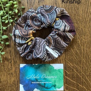 May include: A fabric scrunchie featuring a brown, black, and white dot-patterned design with yellow accents. A small white tag reads Malu Dreamer. Above the scrunchie, the text Kinship is visible. Below, a card displays the text Malu Dreamer.