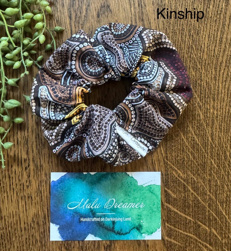 May include: A fabric scrunchie featuring a brown, black, and white dot-patterned design with yellow accents. A small white tag reads Malu Dreamer. Above the scrunchie, the text Kinship is visible. Below, a card displays the text Malu Dreamer.