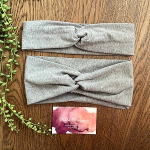 May include: Two grey fabric headbands with a knotted design. The headbands are on a wooden surface with a business card that says "Malu Dreamer Handmade with love. Carefully hand made."