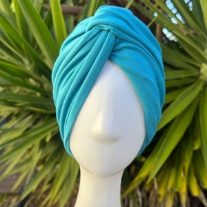 May include: A turquoise turban-style head wrap made of soft fabric. The head covering is displayed on a white mannequin head, with a knotted design at the top. The background features green foliage.