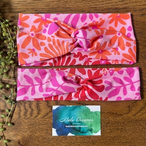 May include: Two knotted headbands with floral patterns in pink, orange, and red. The top headband has a flower design, the bottom one a leaf pattern. A small card with the text "Malu Dreamer" is also visible.