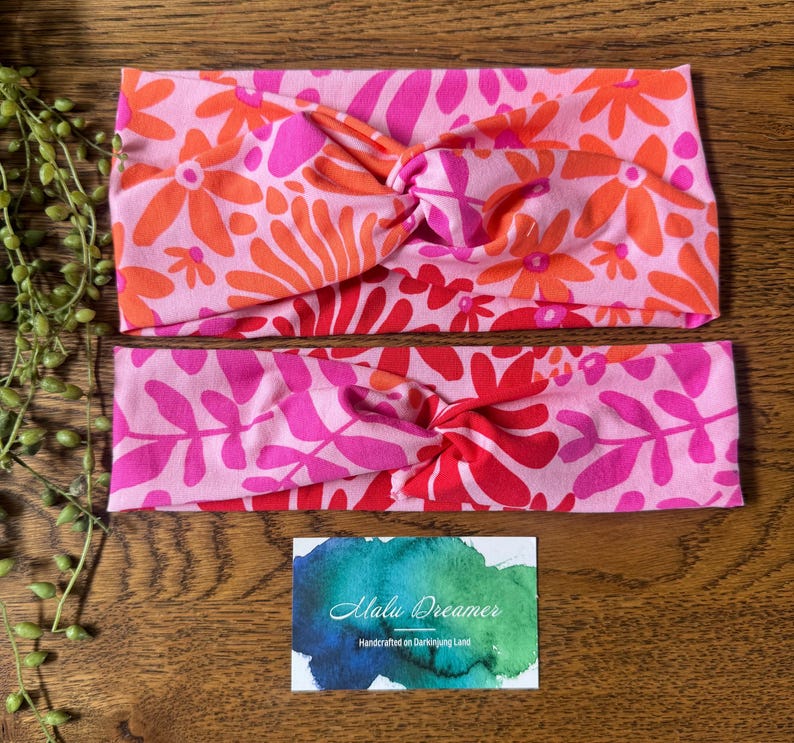 May include: Two knotted headbands with floral patterns in pink, orange, and red. The top headband has a flower design, the bottom one a leaf pattern. A small card with the text "Malu Dreamer" is also visible.