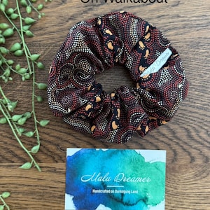 May include: A fabric hair scrunchie featuring a black, red, and orange dot-art pattern. A small white tag is attached to the scrunchie with the text Malu Dreamer. The text On Walkabout is visible above the scrunchie, and a card with the text Malu Dreamer Handcrafted on Darkinjung Land is below.