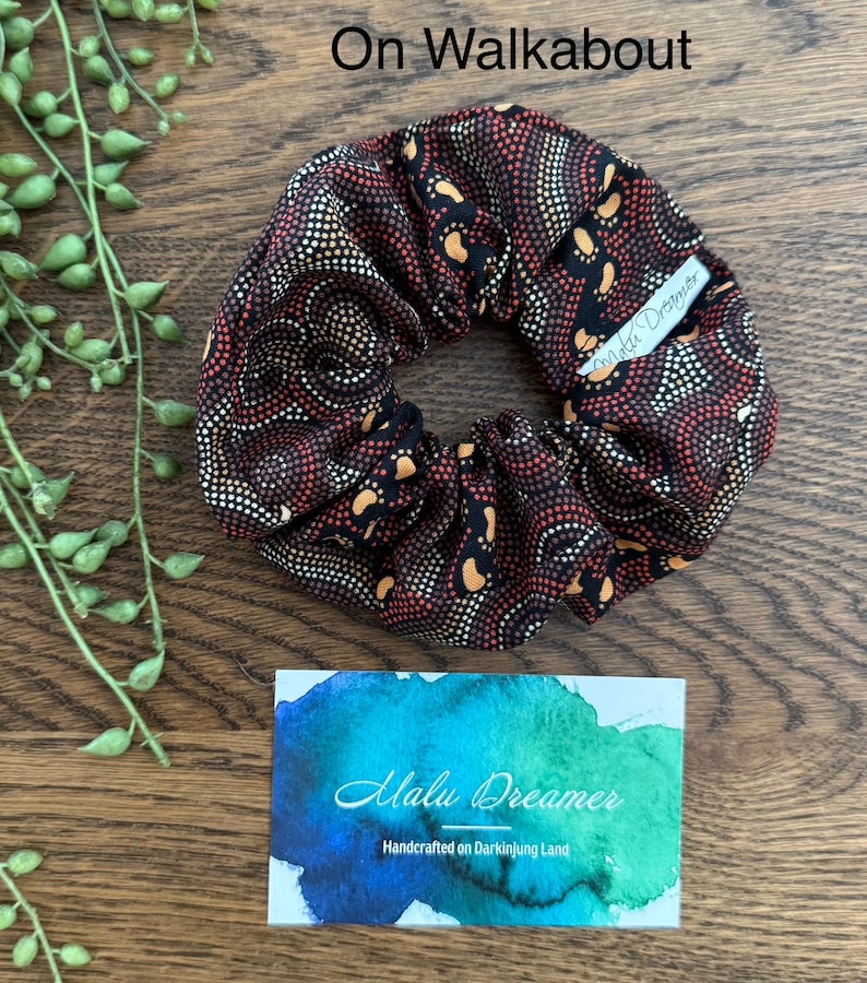 May include: A fabric hair scrunchie featuring a black, red, and orange dot-art pattern. A small white tag is attached to the scrunchie with the text Malu Dreamer. The text On Walkabout is visible above the scrunchie, and a card with the text Malu Dreamer Handcrafted on Darkinjung Land is below.