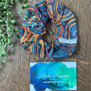 May include: A fabric scrunchie featuring a vibrant, multi-colored pattern of concentric circles and dots in shades of blue, orange, and yellow. A white tag reads Malu Dreamer. A card displays the text Gathering and Malu Dreamer Handcrafted on Darkinjung Land.