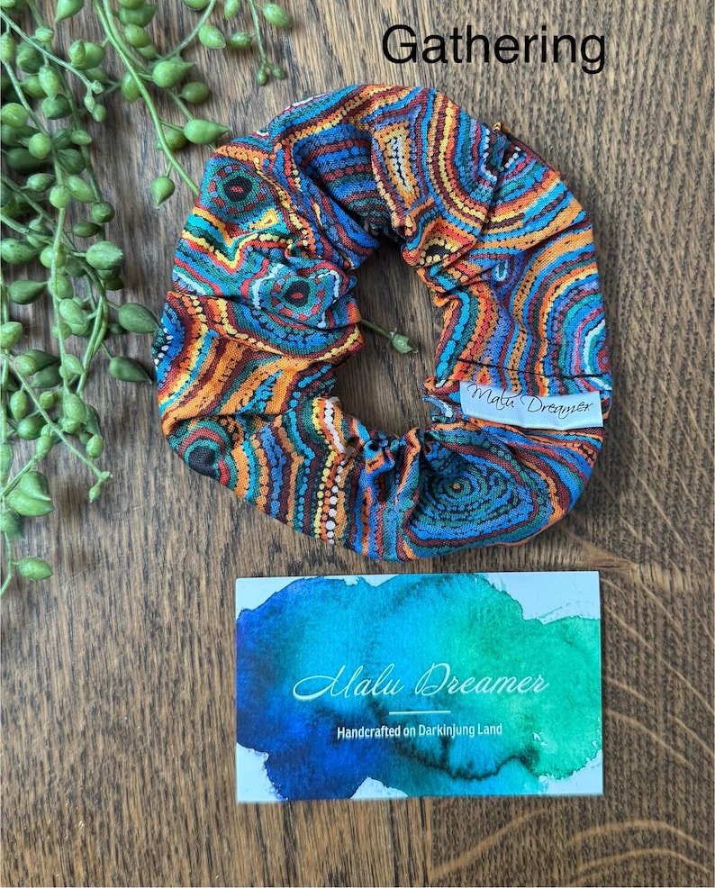 May include: A fabric scrunchie featuring a vibrant, multi-colored pattern of concentric circles and dots in shades of blue, orange, and yellow. A white tag reads Malu Dreamer. A card displays the text Gathering and Malu Dreamer Handcrafted on Darkinjung Land.