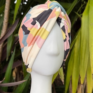 May include: A fabric turban headband featuring a knotted front detail and a multi-colored abstract pattern in shades of cream, black, soft pink, yellow, and muted blue, displayed on a white mannequin head to show the wrap-around design.
