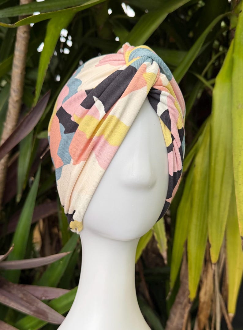 May include: A fabric turban headband featuring a knotted front detail and a multi-colored abstract pattern in shades of cream, black, soft pink, yellow, and muted blue, displayed on a white mannequin head to show the wrap-around design.