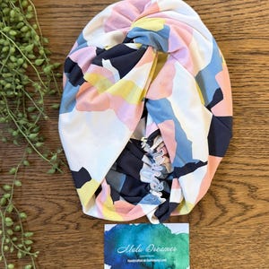 May include: A fabric headwrap featuring a twisted knot design with an abstract pattern of white, navy blue, soft pink, and yellow shapes. The headwrap includes an elasticized back section and is displayed with a card labeled Malu Dreamer.