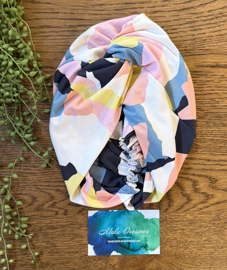 May include: A fabric headwrap featuring a twisted knot design with an abstract pattern of white, navy blue, soft pink, and yellow shapes. The headwrap includes an elasticized back section and is displayed with a card labeled Malu Dreamer.