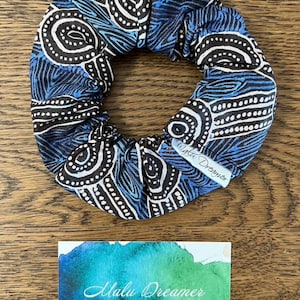 May include: A fabric hair scrunchie featuring a blue, black, and white pattern with swirling lines and dotted circles. A white fabric tag attached to the scrunchie reads Malu Dreamer. Below is a card with a blue and green watercolour design.