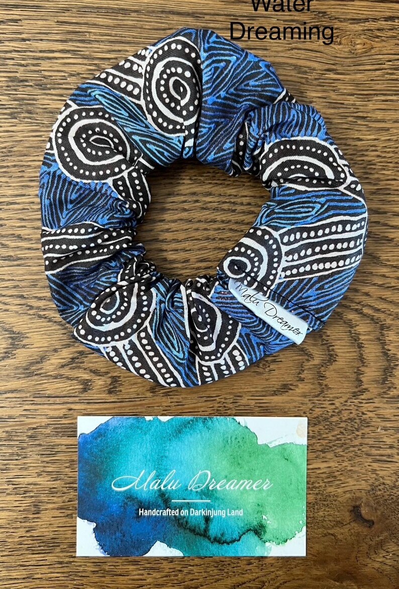 May include: A fabric hair scrunchie featuring a blue, black, and white pattern with swirling lines and dotted circles. A white fabric tag attached to the scrunchie reads Malu Dreamer. Below is a card with a blue and green watercolour design.