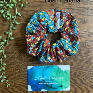 May include: A fabric hair scrunchie featuring a repeating pattern of small yellow, blue, and red flowers. A white tag is attached to the scrunchie. The text Bush banana is visible above, and a card with the text Malu Dreamer is below.